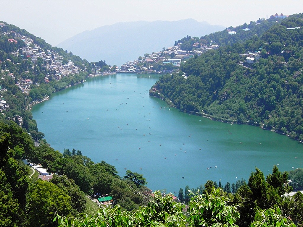 A scenic view of Naini Lake surrounded by lush green hills and hillside buildings in Nainital, India.