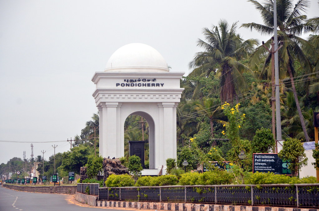A grand white gateway with a dome marks the entrance to Pondicherry, surrounded by palm trees and greenery.