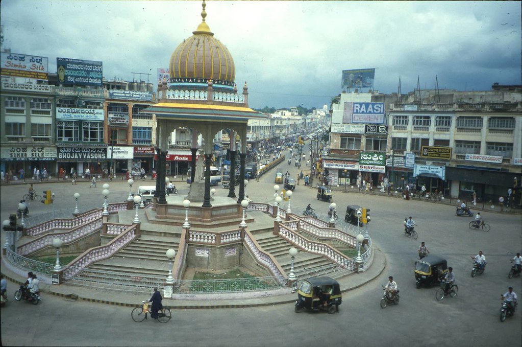 A busy intersection in Mysore featuring the iconic domed pavilion, surrounded by historic commercial buildings and lively street activity.